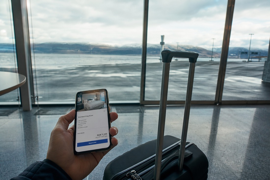 Traveller's phone screen showing a hotel booking beside a rolling suitcase in a bright airport arrivals hall