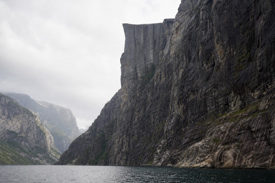 Sheer cliff face rising from a narrow fjord seen from water level with a flat rock shelf high above