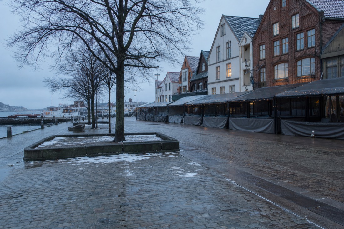 Empty snow-dusted Norwegian city square with closed cafe awnings and bare trees in winter light