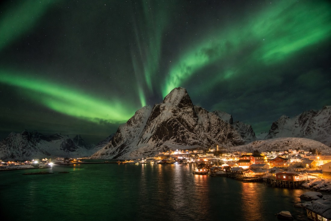 Aurora over a snow-covered fjord village with lit windows and mountains behind