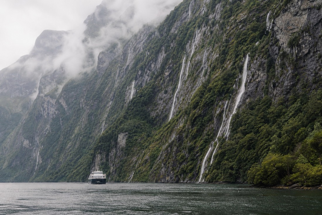 Fjord cruise boat seen from the shore against tall green cliffs with waterfalls
