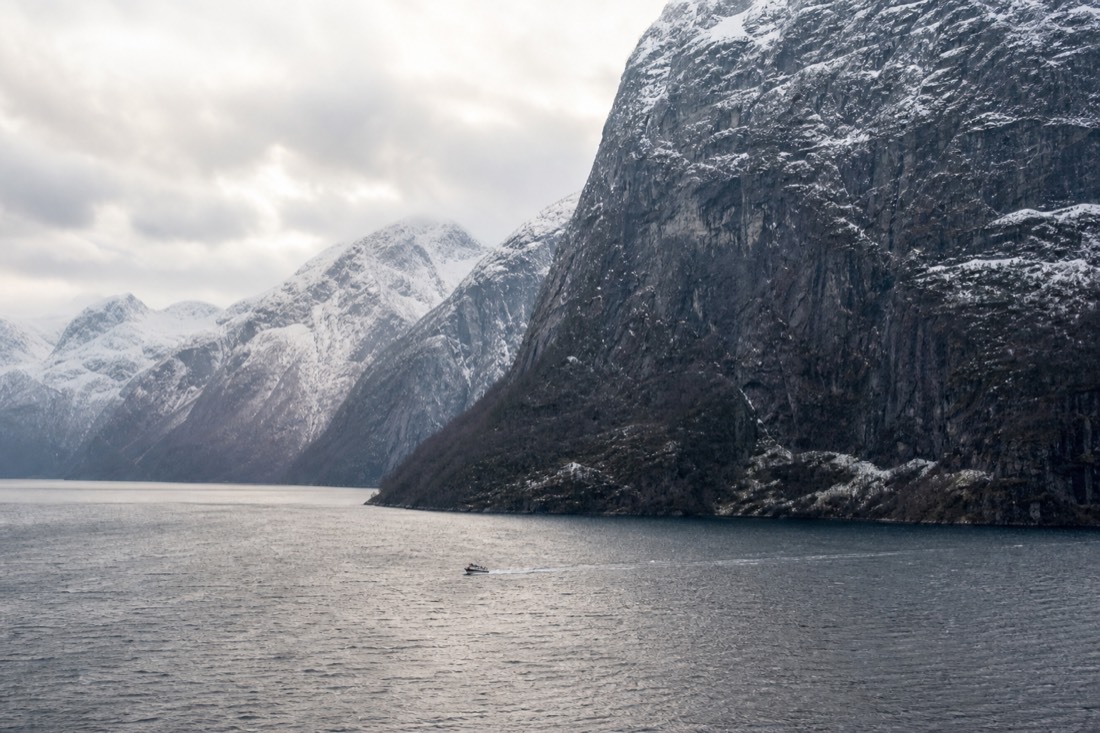 Snow-dusted fjord walls above dark water with a small boat leaving a wake