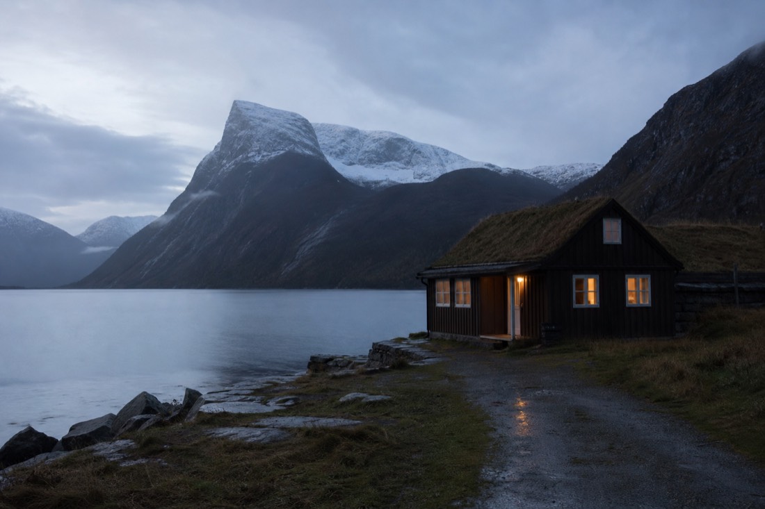Small wooden guesthouse beside a fjord with a few windows lit at dusk