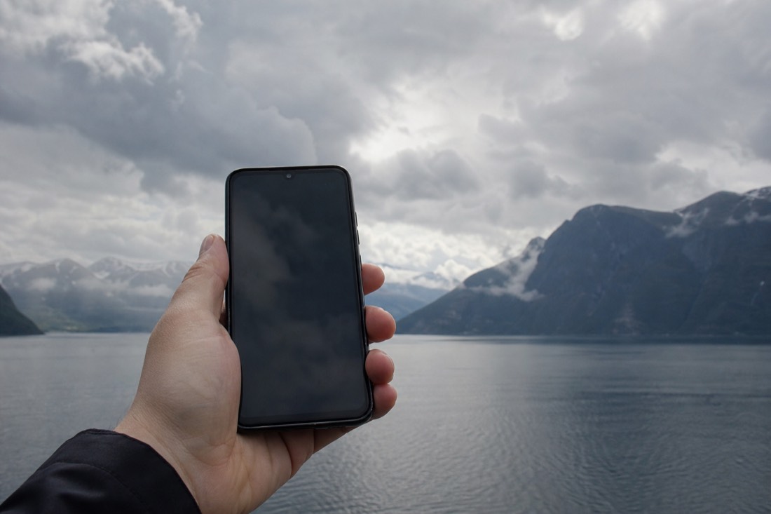 Phone showing a blank weather app screen held up against a cloudy fjord sky