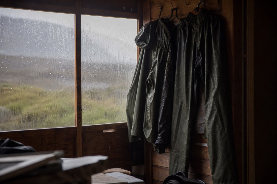 Waterproof jacket and over-trousers hanging to dry beside a window streaked with rain