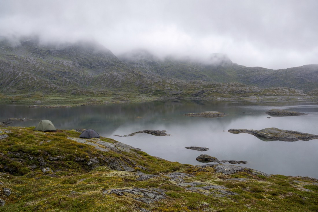 Two small tents on a mossy plateau beside a still mountain lake with cloud sitting low on the ridge behind