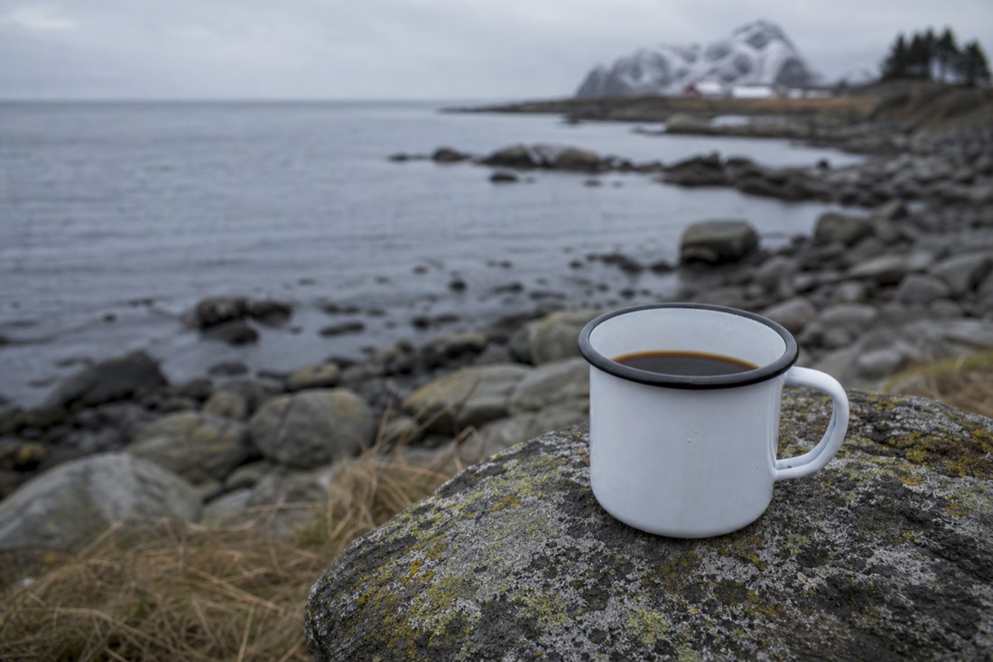 Enamel mug of black coffee resting on a rock outdoors with cold water behind