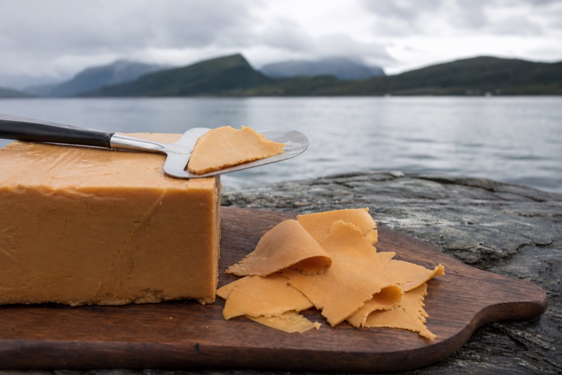 Block of Norwegian brown cheese with a cheese slicer and thin curled slices on a wooden board