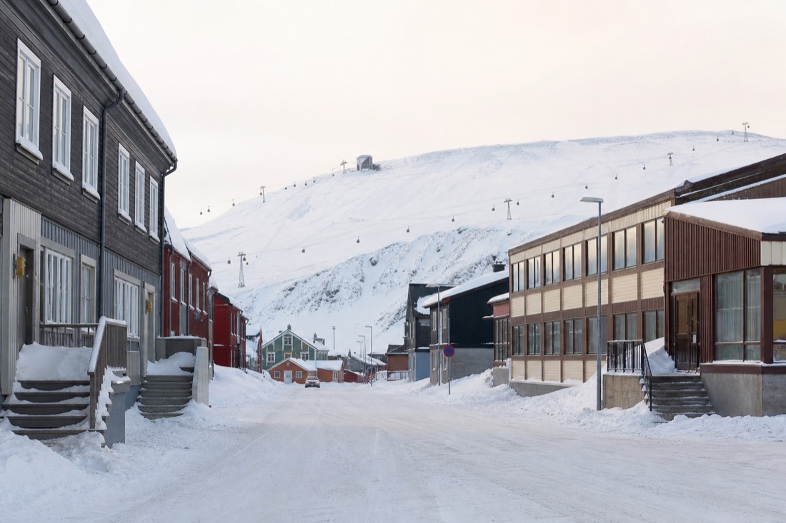 Snowy Arctic town street in daylight with shops and a cable car line visible on the hill behind