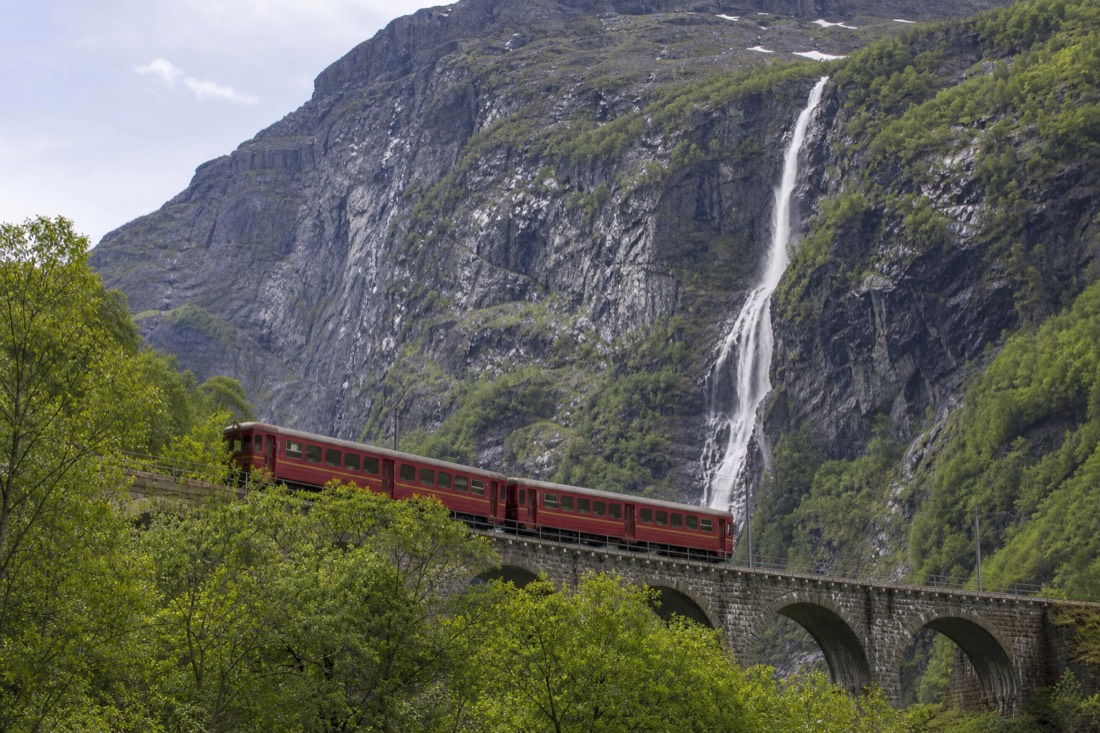 A red mountain railway carriage crossing a viaduct above a green Norwegian valley with a waterfall behind