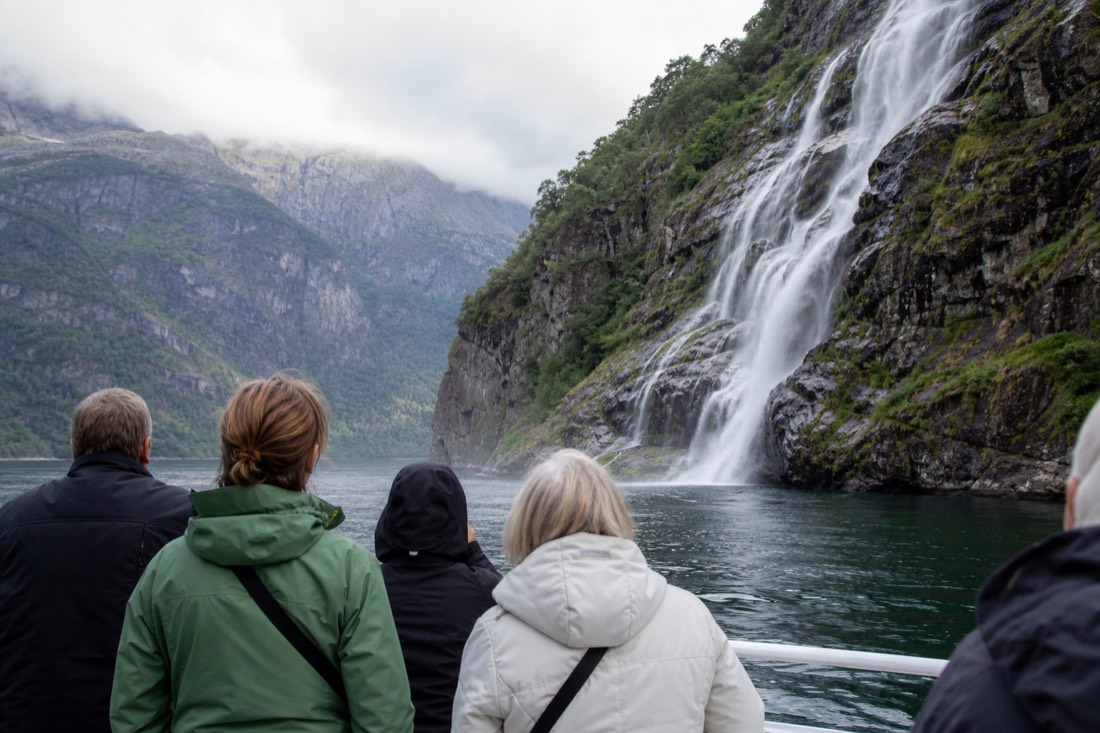 Passengers on an open boat deck watching a waterfall drop into narrow fjord water near Bergen
