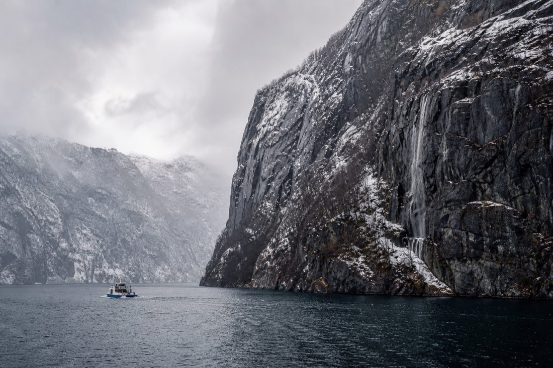 Snow-dusted fjord walls rising above dark winter water with a small boat below