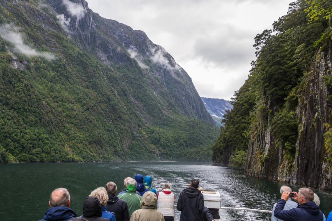 Passengers on an open boat deck looking up at steep green fjord walls close on both sides