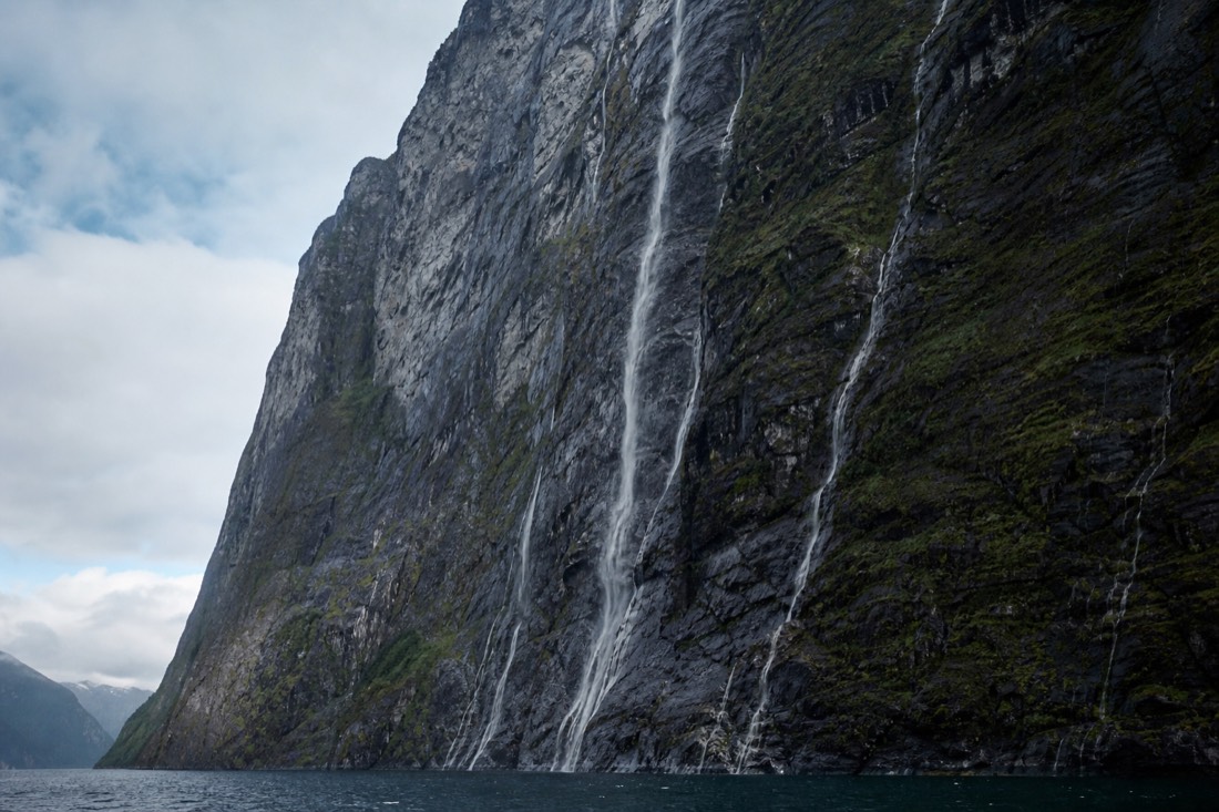 View up at a sheer cliff face from a boat on a narrow fjord with waterfalls on the rock