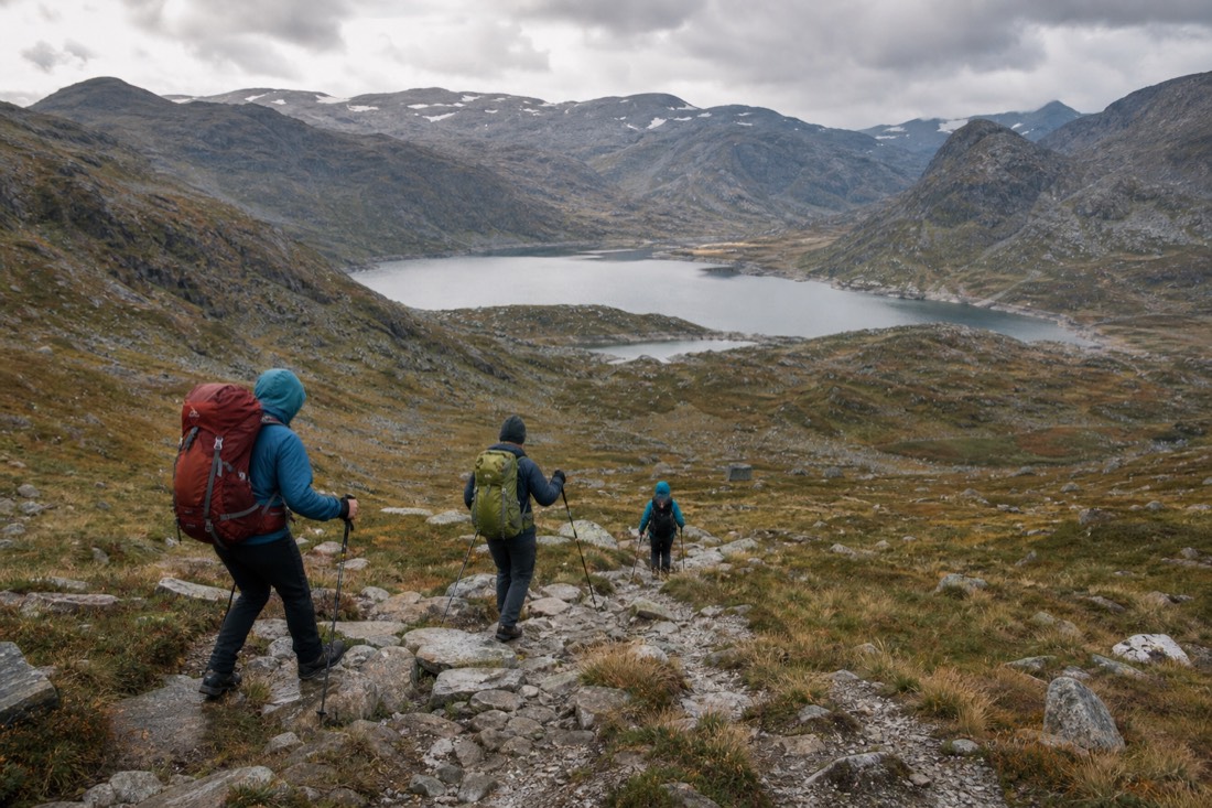 Walkers descending a rocky Norwegian trail with poles, mountains and a lake behind them