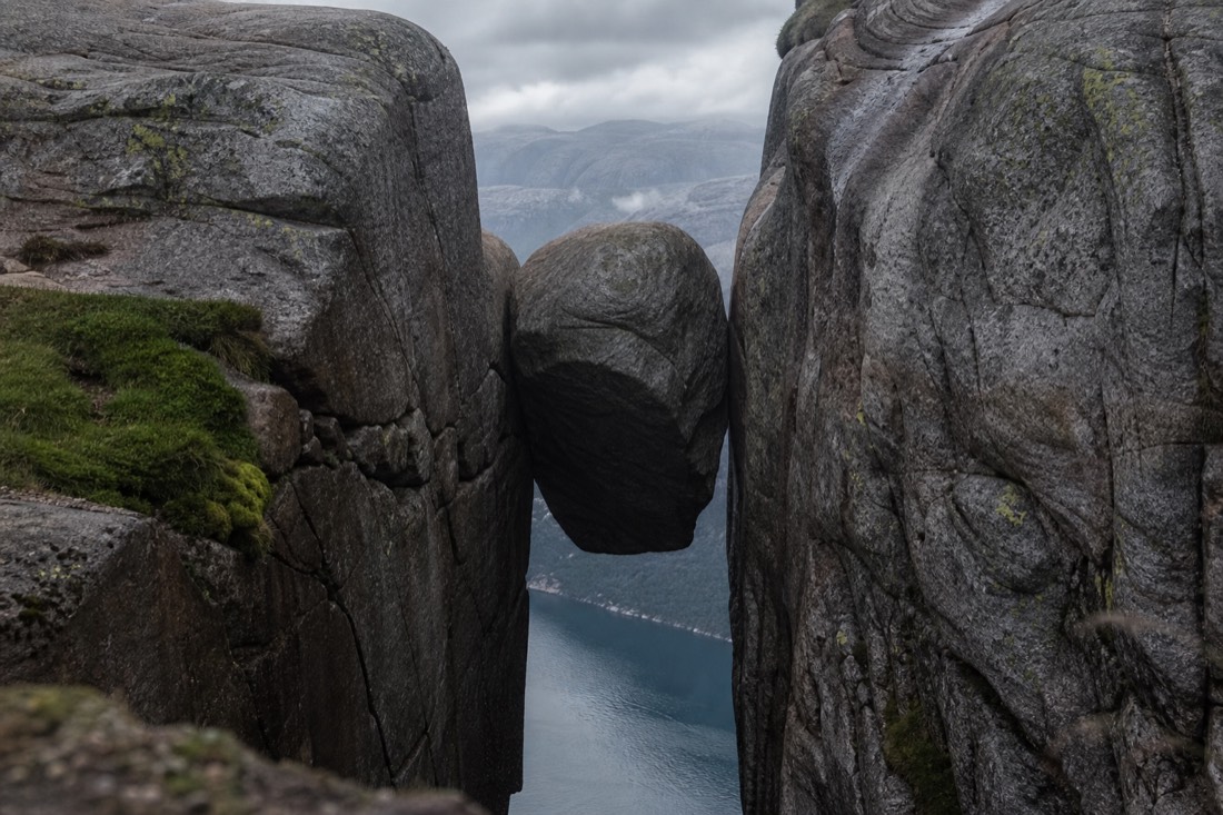 Round boulder wedged between two vertical rock faces high above a narrow fjord in cloudy weather