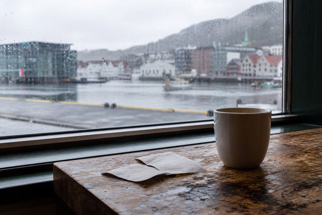 Two supermarket receipts and a coffee cup on a wooden cafe table beside a harbour window in Bergen