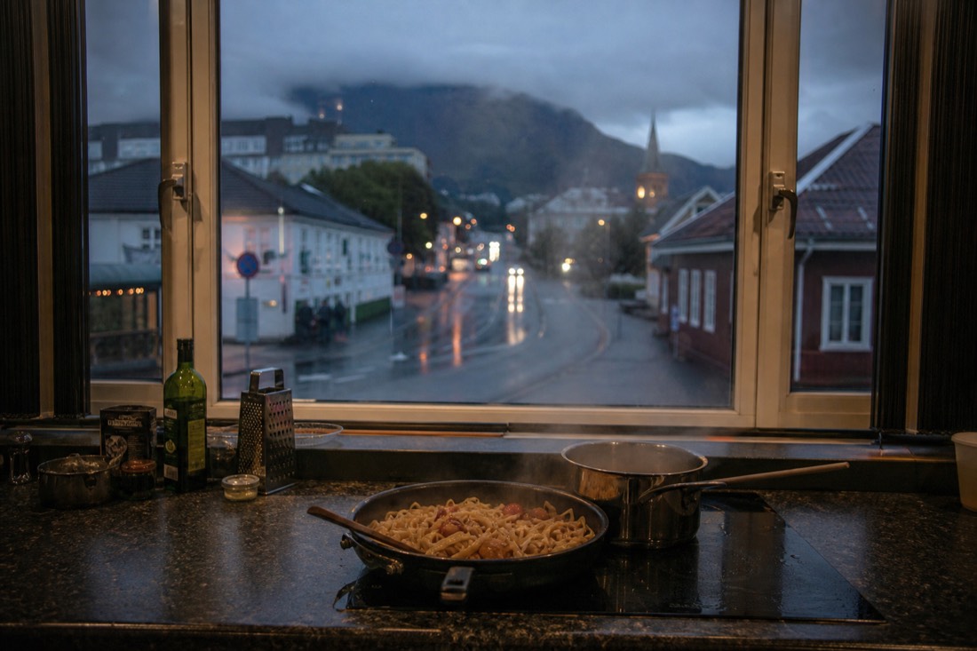Hostel kitchen counter with a simple pasta dinner and a window looking onto a wet Norwegian street at dusk