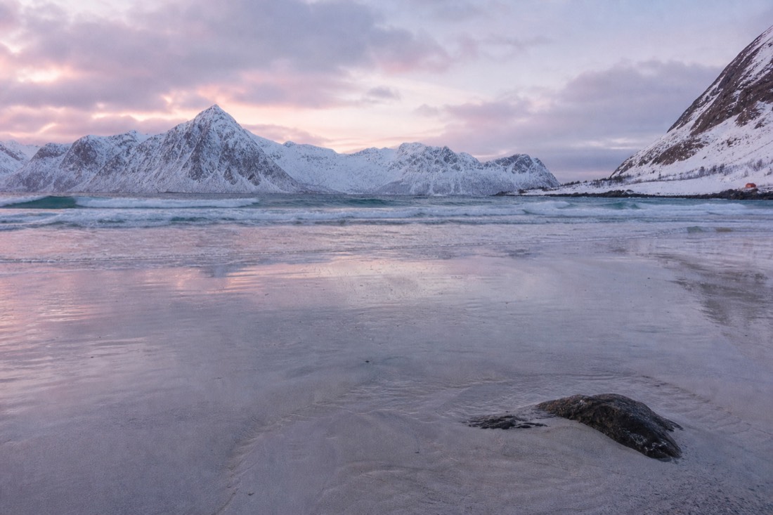 Winter beach in Lofoten with pale sand, dark water and snow-covered peaks under low light