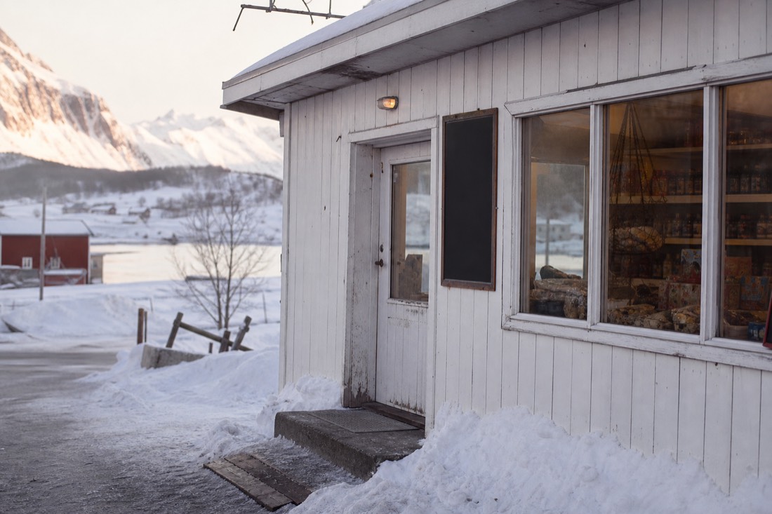 Small village grocery shop front in Lofoten with a hand-lettered opening hours sign and snow banked outside