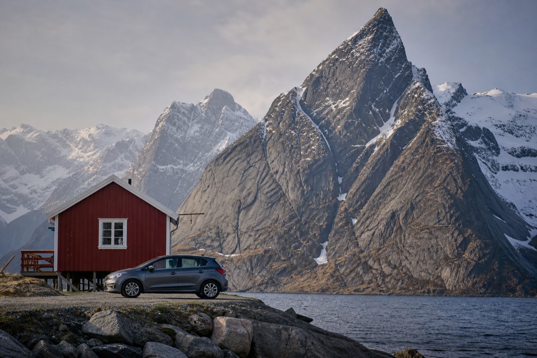 Rental car parked beside a red rorbuer cabin at the water's edge with granite peaks rising behind