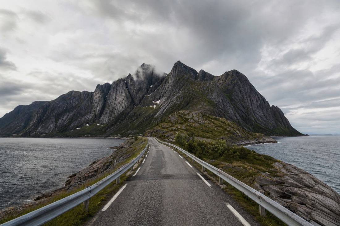 Empty single-lane road running between steep Lofoten peaks with the sea visible on both sides