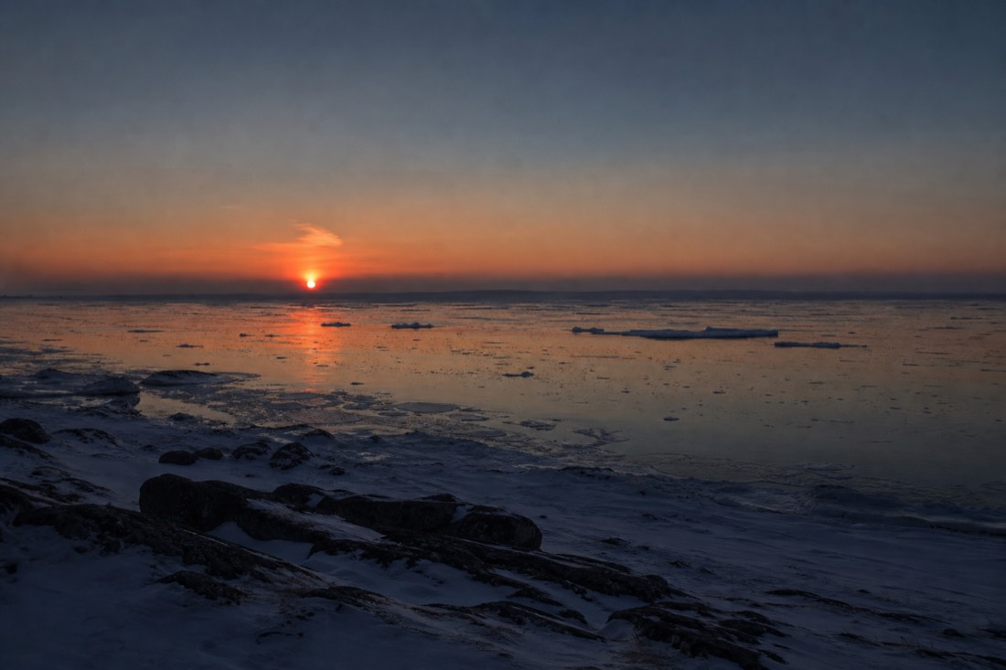 The sun sitting just above the horizon at night over an Arctic coastline with orange light on the water