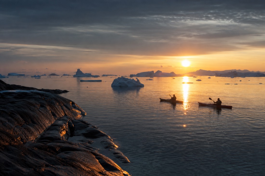 Two kayakers on glassy Arctic water at midnight with a low sun casting a long copper reflection