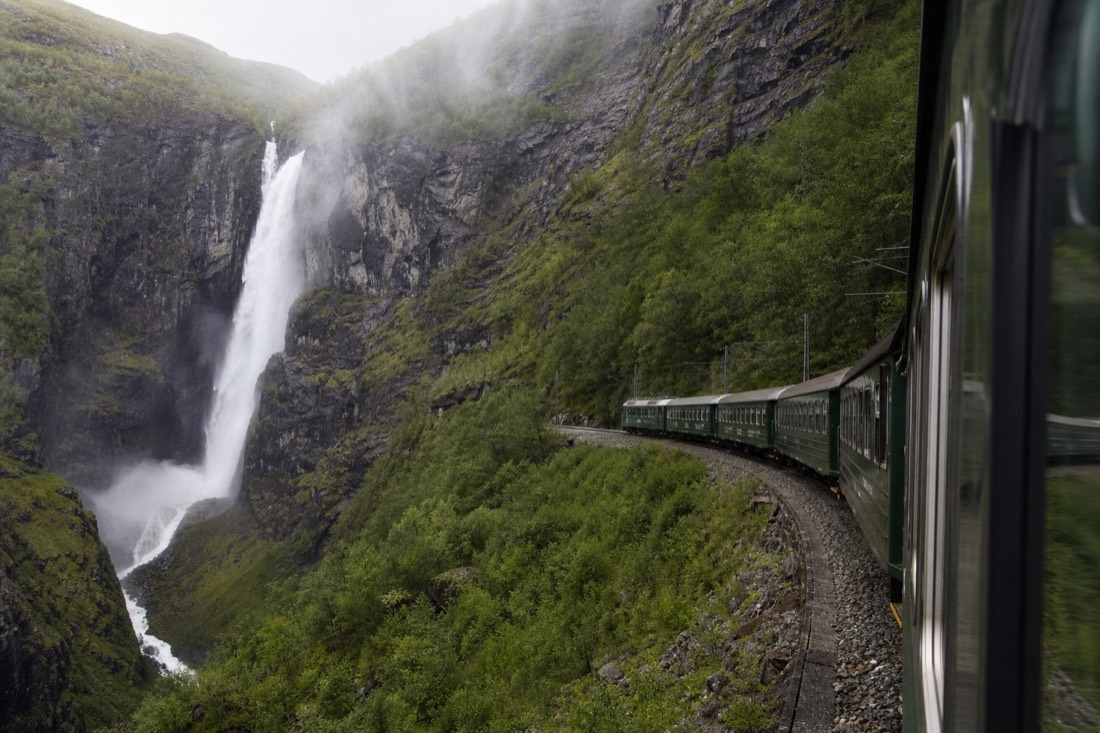 Green railway carriages on a steep mountainside track beside a waterfall in a narrow valley