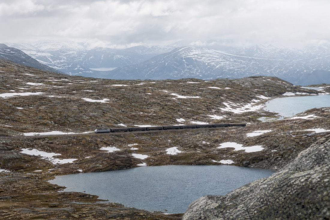 Mainline train crossing a high bare mountain plateau with patches of snow and small lakes