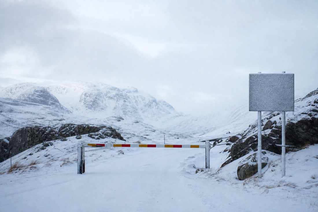 Closed road barrier across a snow-covered mountain pass with a seasonal closure sign post beside it
