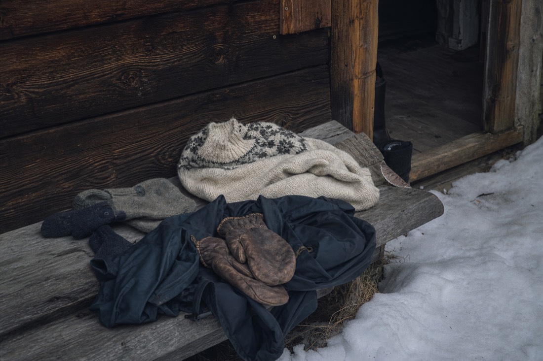 Wool layers, a windproof shell and thick mittens laid out on a wooden bench by a snowy doorway