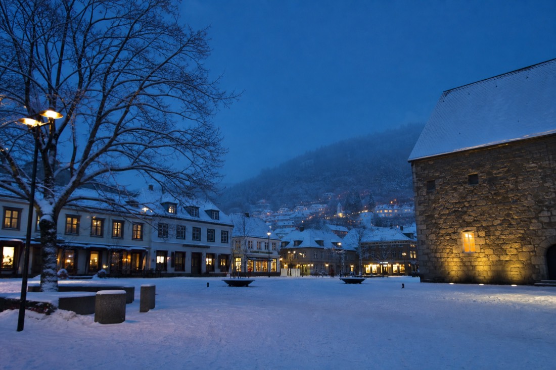 Snowy Norwegian town square at midday in December with blue light and warmly lit shop fronts