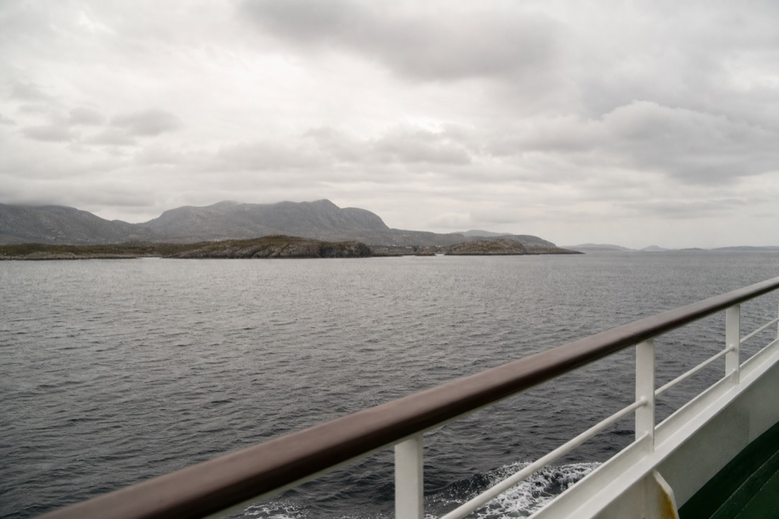 Ferry deck railing with the Norwegian coastline and low islands passing under grey cloud