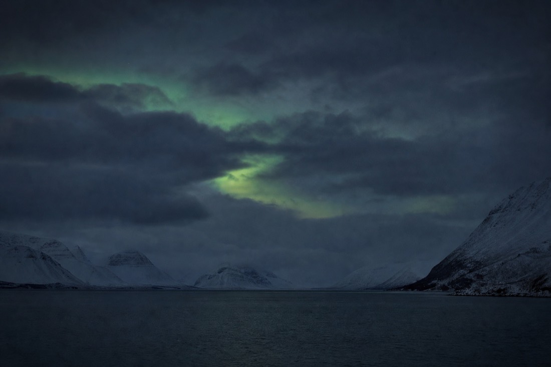 Cloudy Arctic night sky over a fjord with a thin break in the cloud and faint green light showing