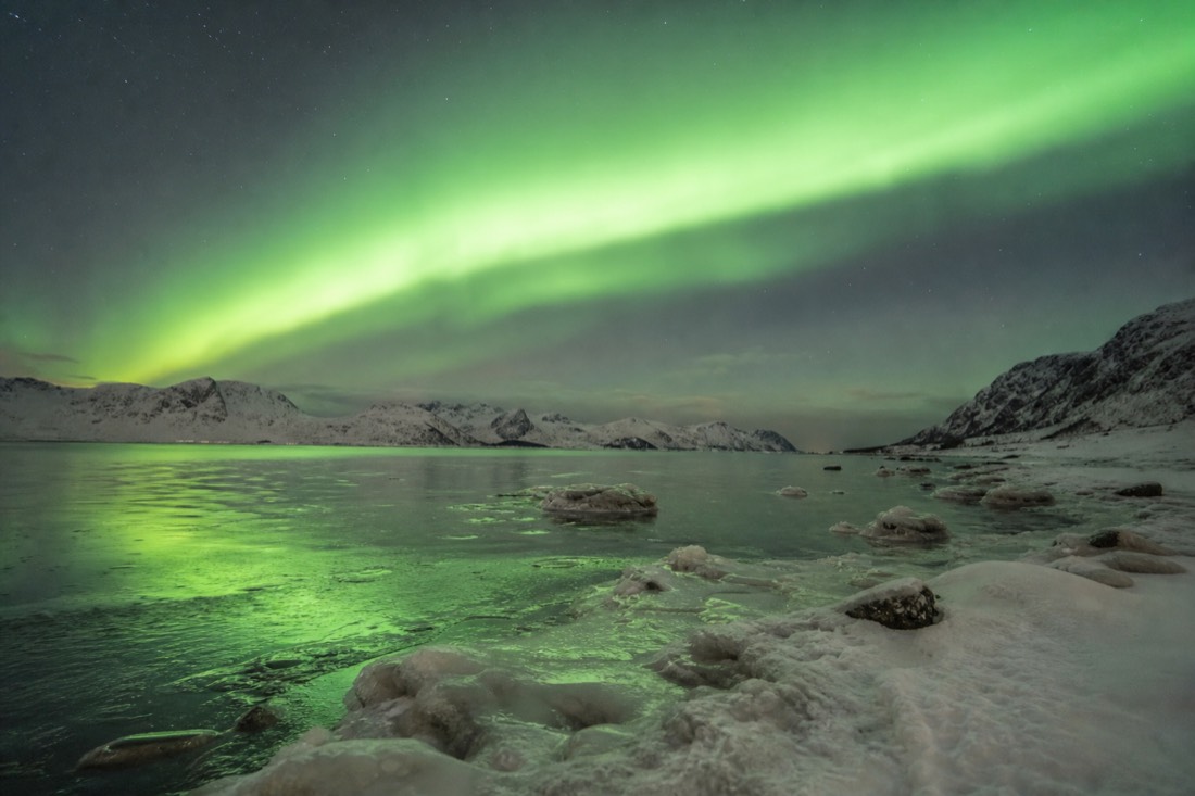 Aurora arc over a snow-covered Arctic landscape with a frozen shoreline in the foreground
