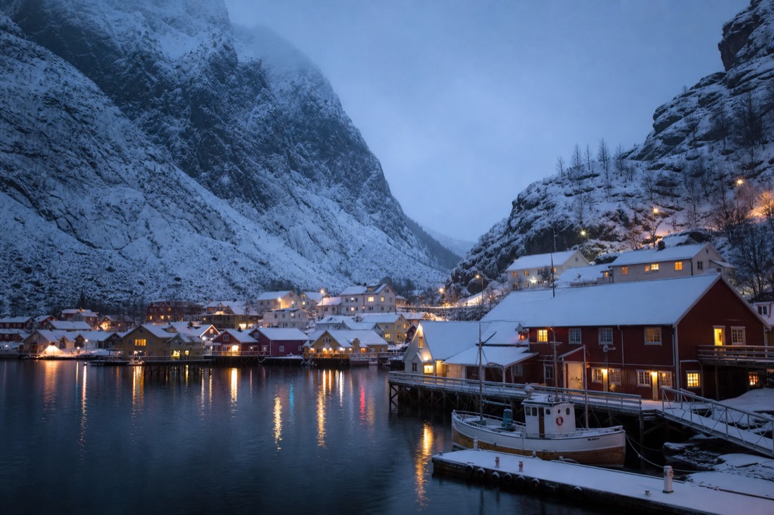 Snow-covered fjord village at dusk with lit windows and a boat moored at a quiet jetty