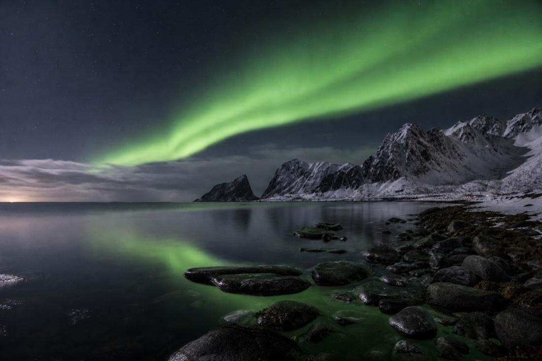 Green aurora arcing over a snow-covered Arctic shoreline with dark mountains reflected in still water