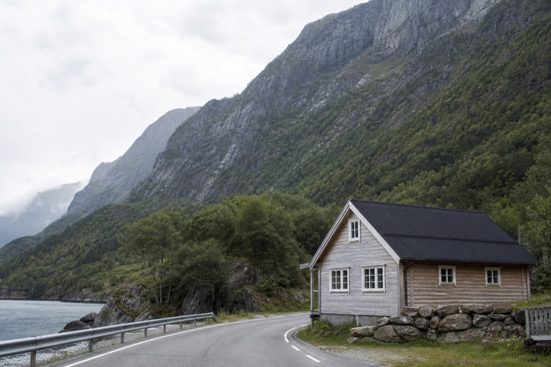 Small wooden guesthouse beside a quiet fjord road with mountains rising directly behind it