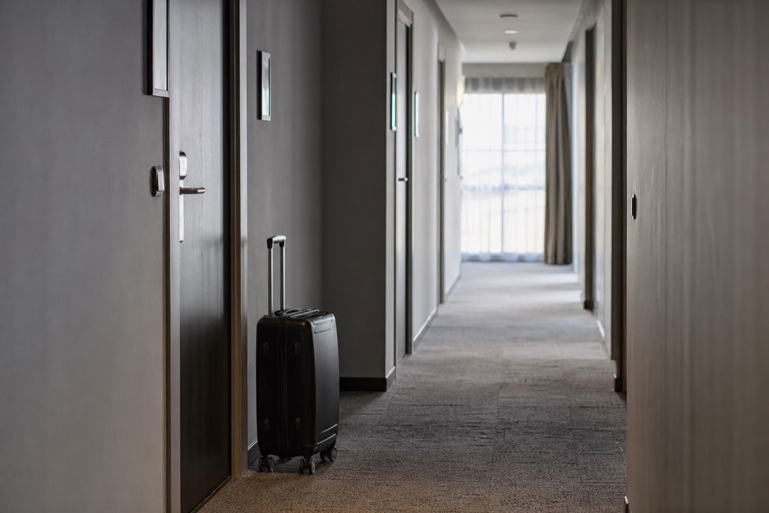 Hotel corridor with a wheeled suitcase beside a door and soft daylight from a window at the end