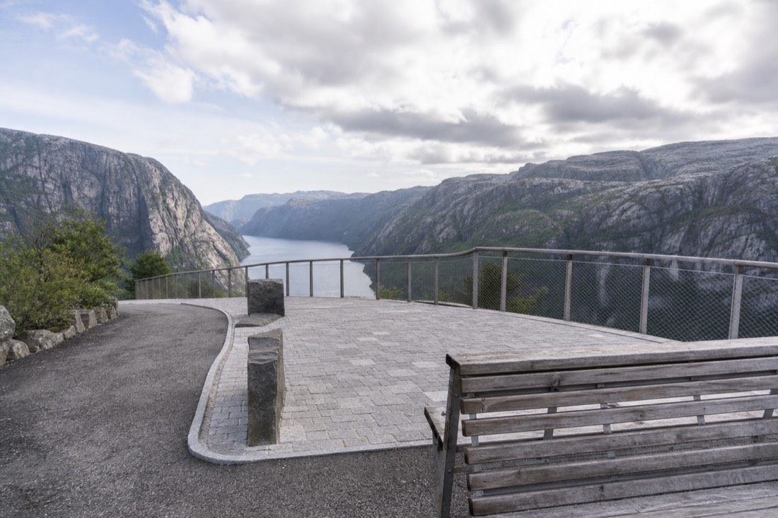 Wide viewing platform with a railing looking down a fjord, a bench in the foreground and gentle paved approach