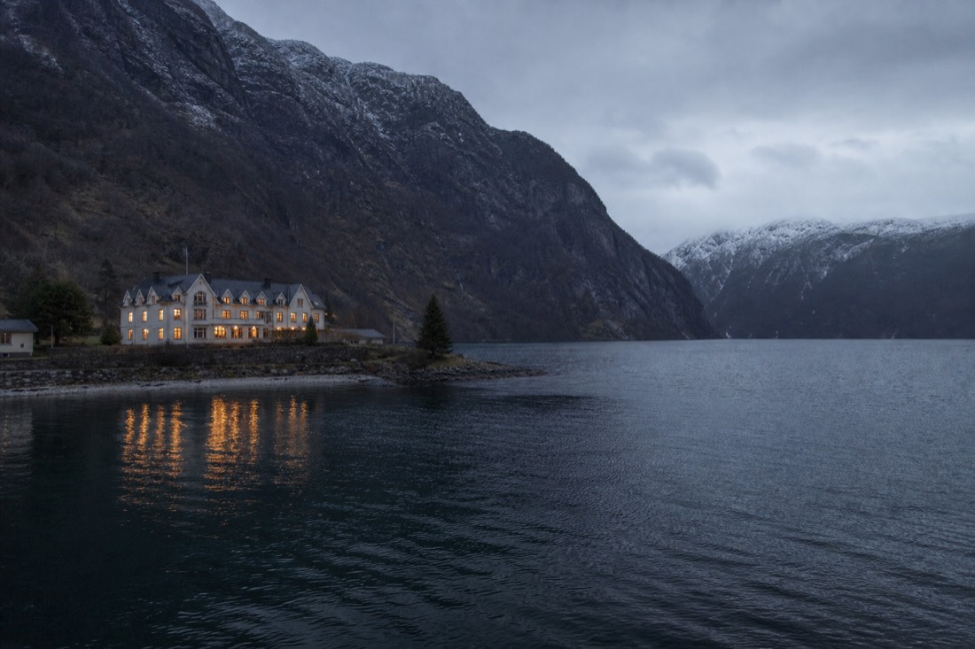 Small hotel with lit windows beside a dark fjord in early evening with snow on the far ridge