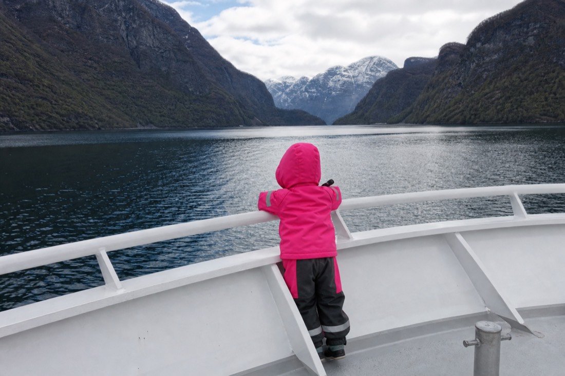 Child in a bright snowsuit watching from the deck rail of a small boat in a calm fjord