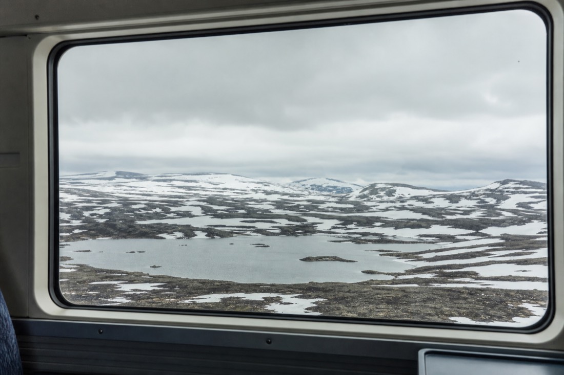 Train window looking out over a snow-patched high plateau with small lakes and no trees