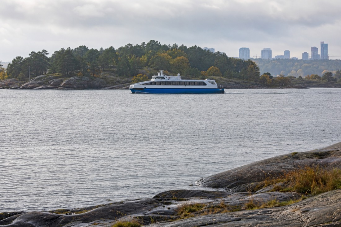 Electric passenger catamaran gliding past low wooded islands on calm water near a city skyline