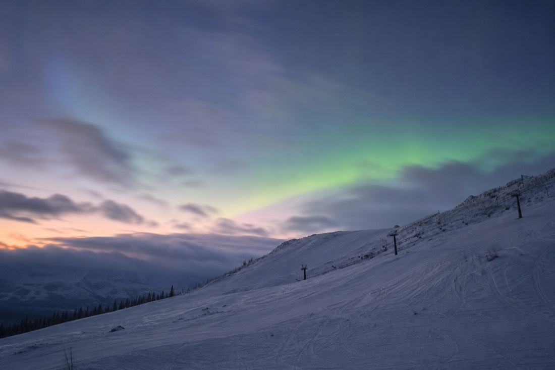 Snowy slope at dusk with a faint green glow beginning in the sky above the treeline