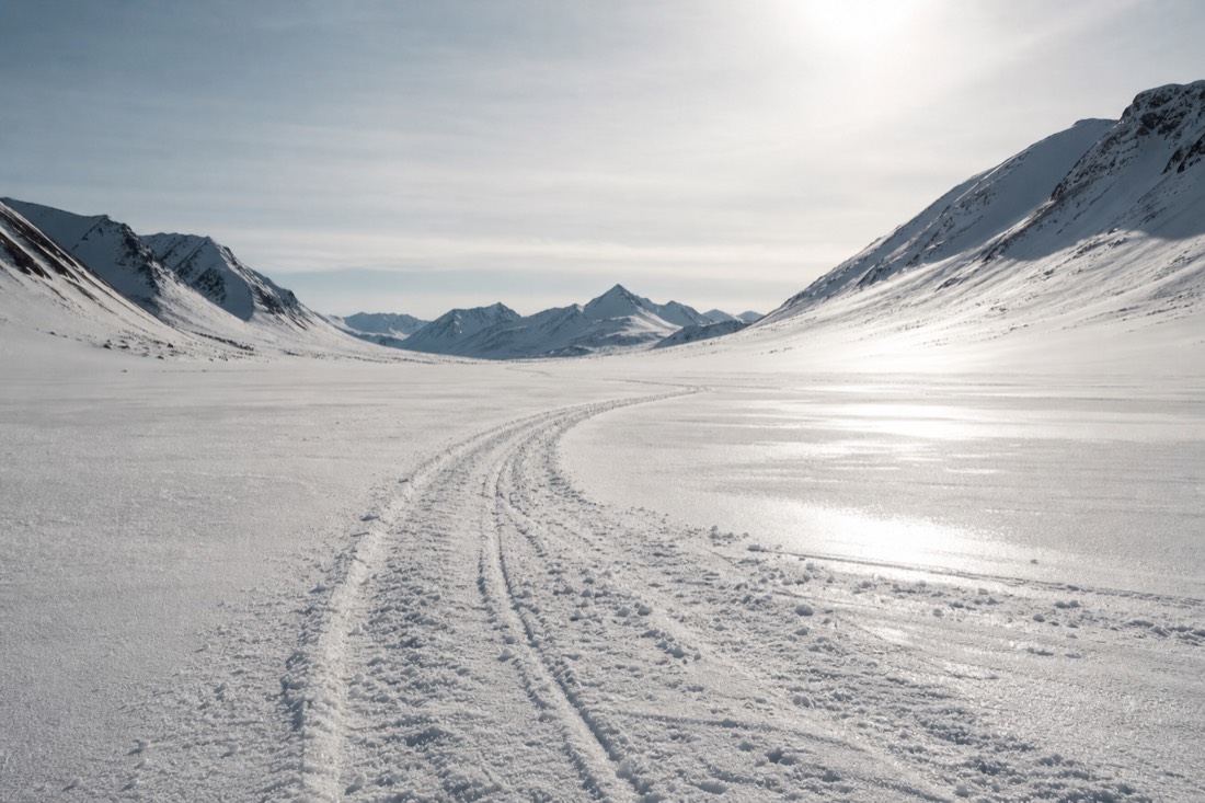 Snowmobile tracks crossing a wide white Arctic valley under low bright spring light