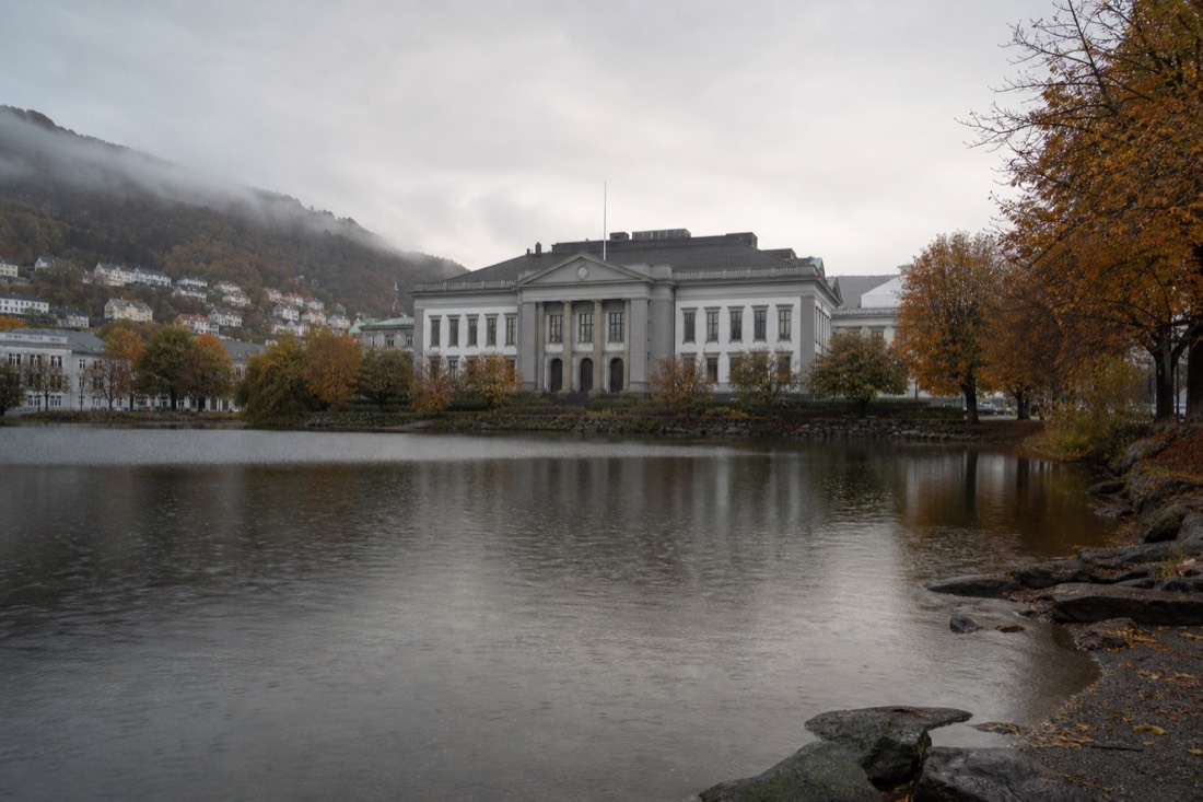 Neoclassical museum building beside a still city lake in Bergen with autumn trees along the shore