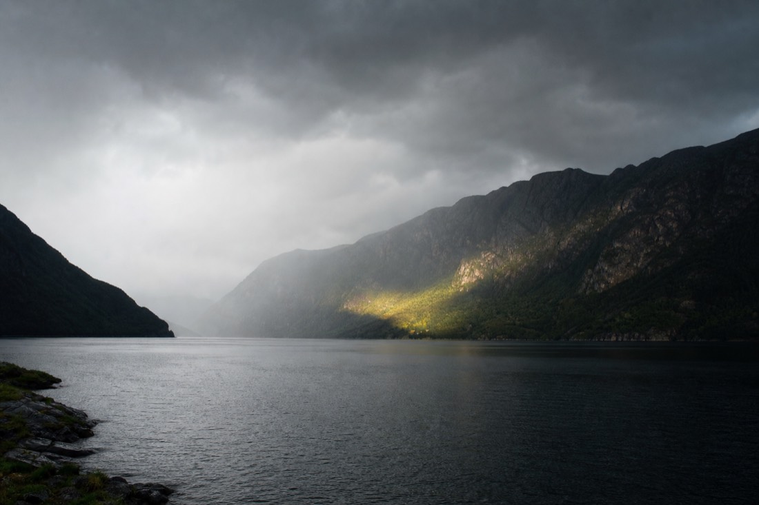 Dark weather front moving across a fjord with sunlight breaking through on the far shore
