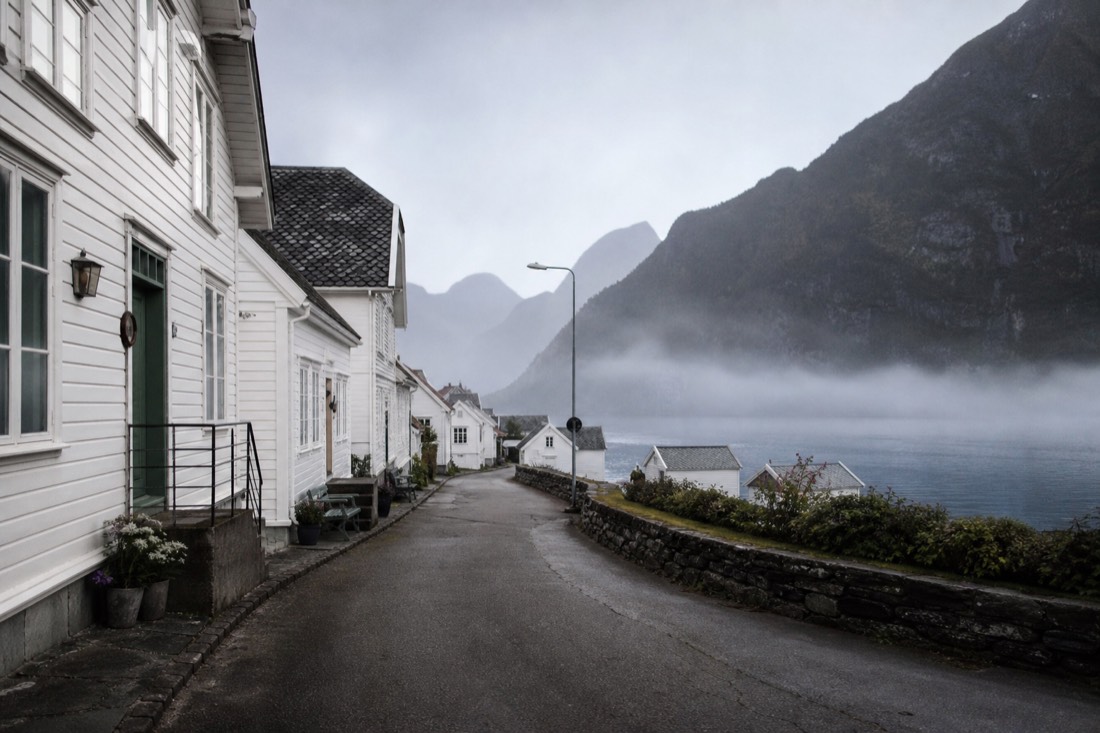 Empty village street beside a fjord in early morning with mist on the water and closed shutters